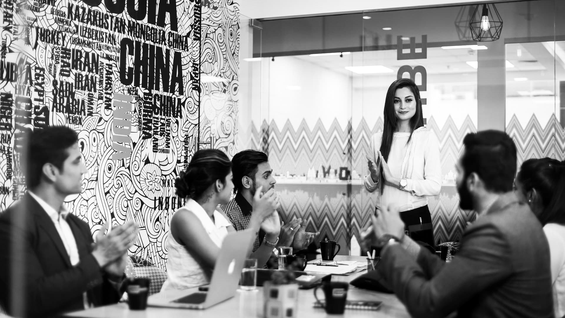 An office meeting: a woman stands at the head of the table and presents while her colleagues listen and applaud. Black and white.